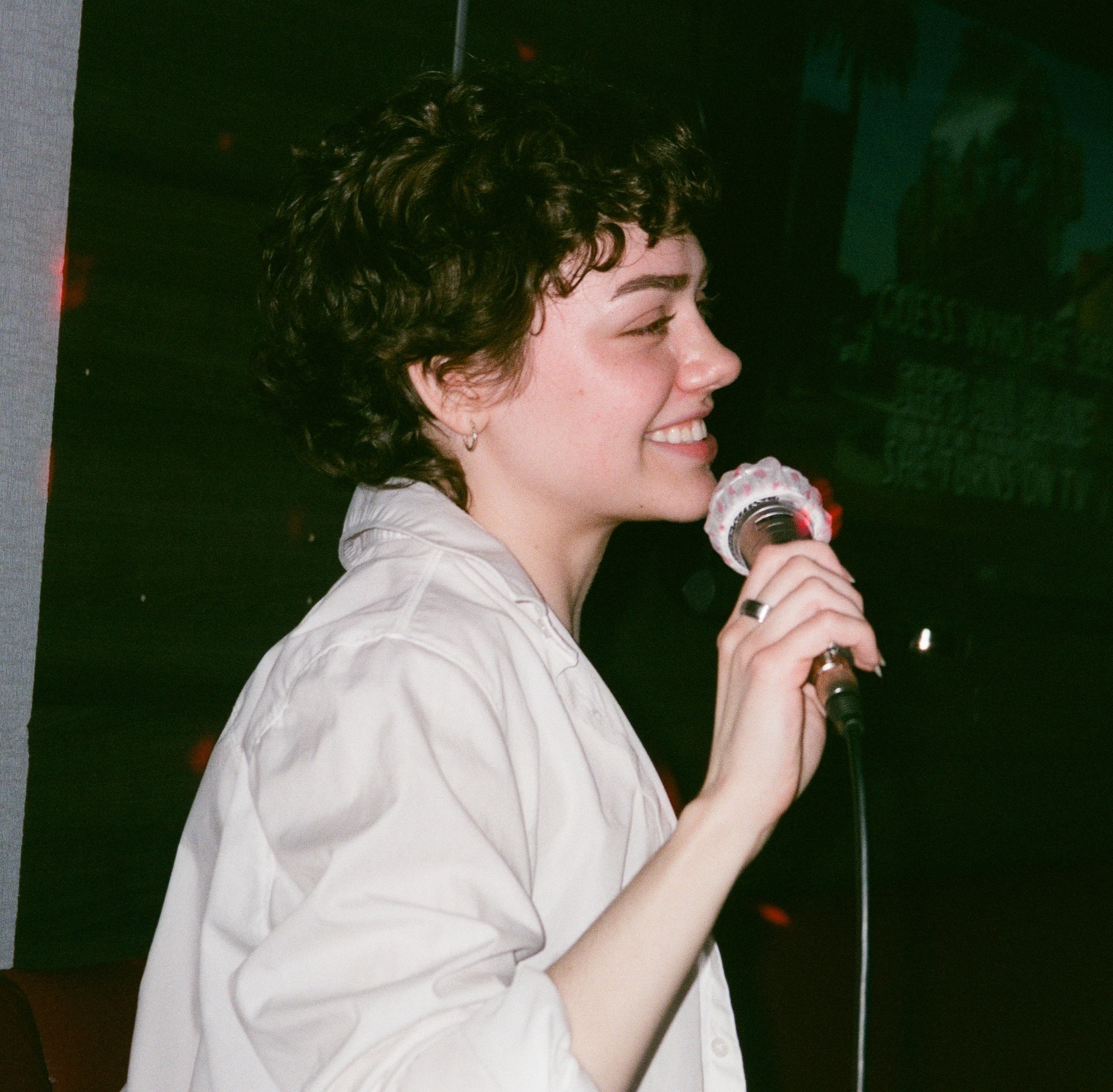 A photo of Lily Mackillican holding a karaoke microphone and smiling. They have brown curly hair, pale skin, and are wearing a white shirt.